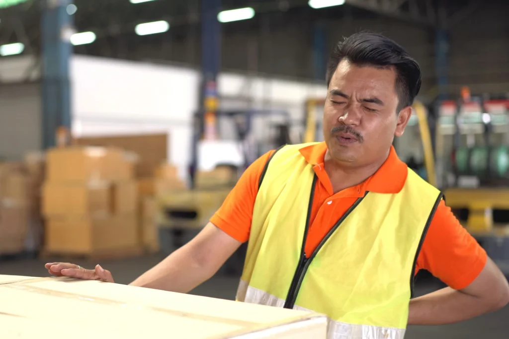 A warehouse worker in a hi-vis vest pausing with a hand to a sore back